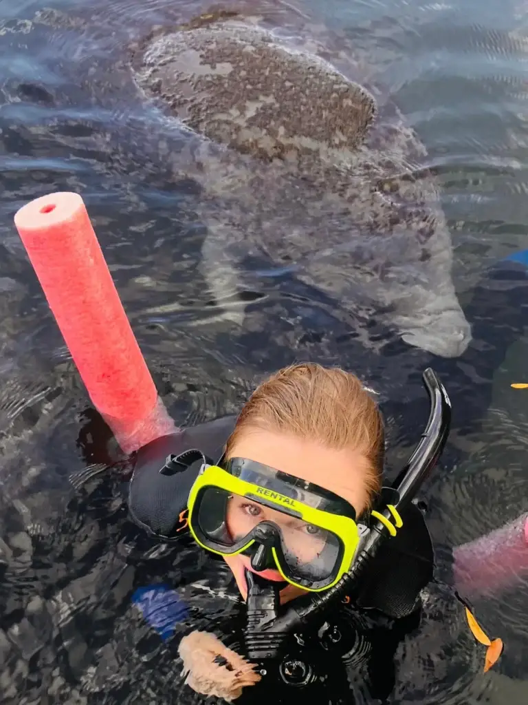 Smiling snorkeler in a yellow mask with a manatee swimming behind her